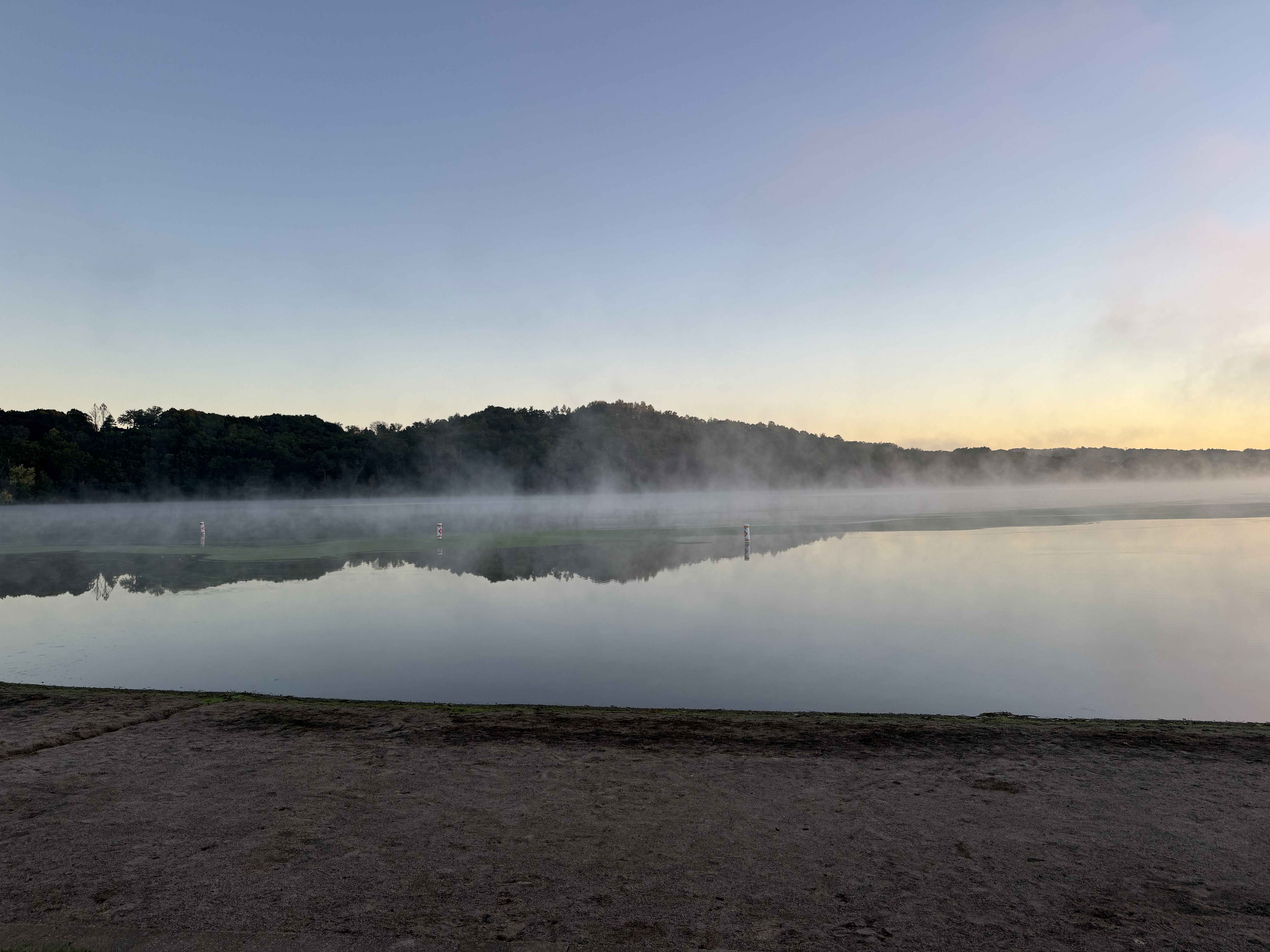 A tranquil lakeside scene at dawn, featuring gentle mist rising from the water’s surface, with a calm reflection of trees and the sky. The shoreline is sandy, and faint outlined markers can be seen in the water.
