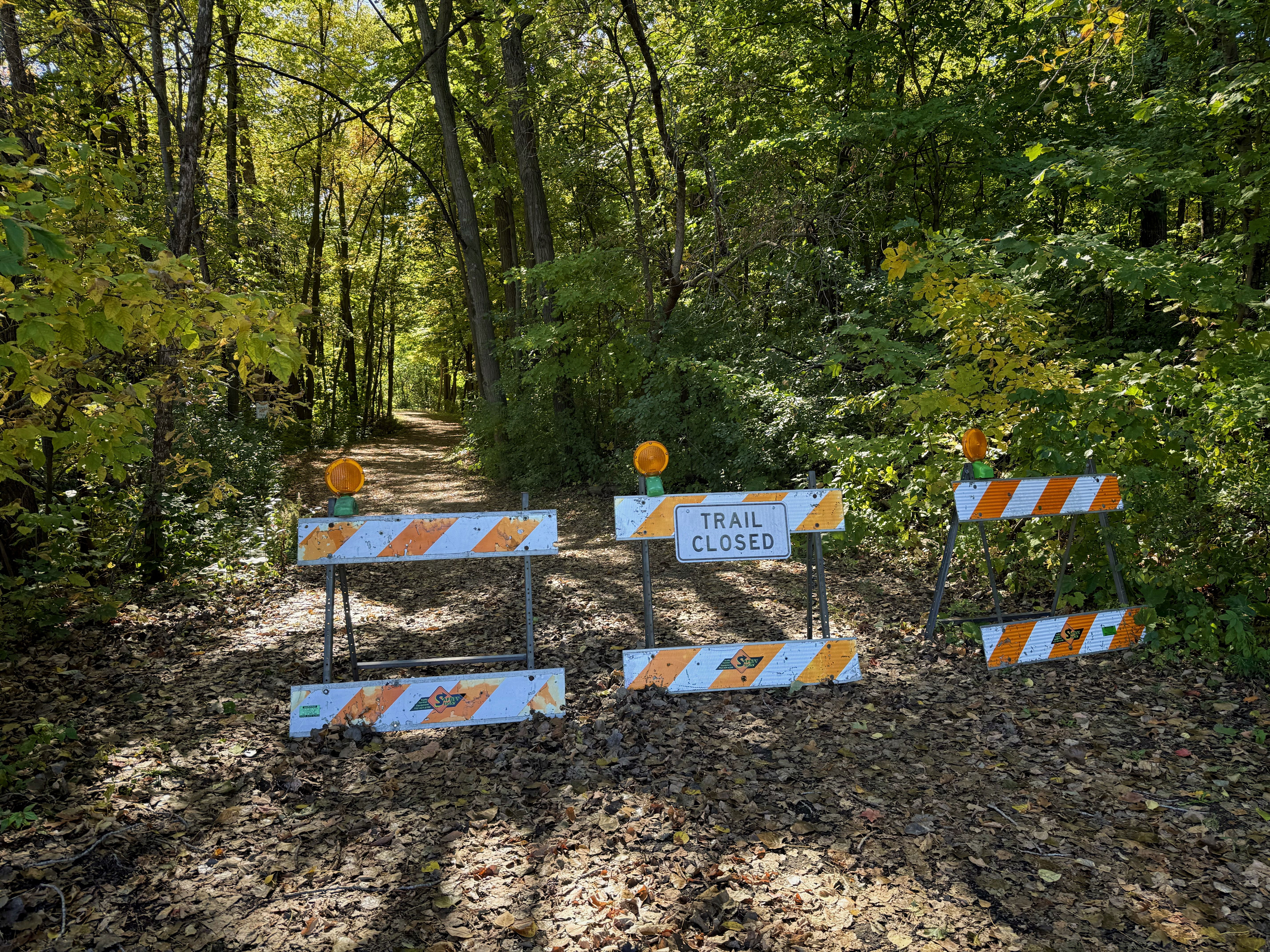 A trail closed sign with barriers is positioned across a dirt path surrounded by trees with green and yellow leaves, indicating it’s autumn. The ground is covered with fallen leaves.