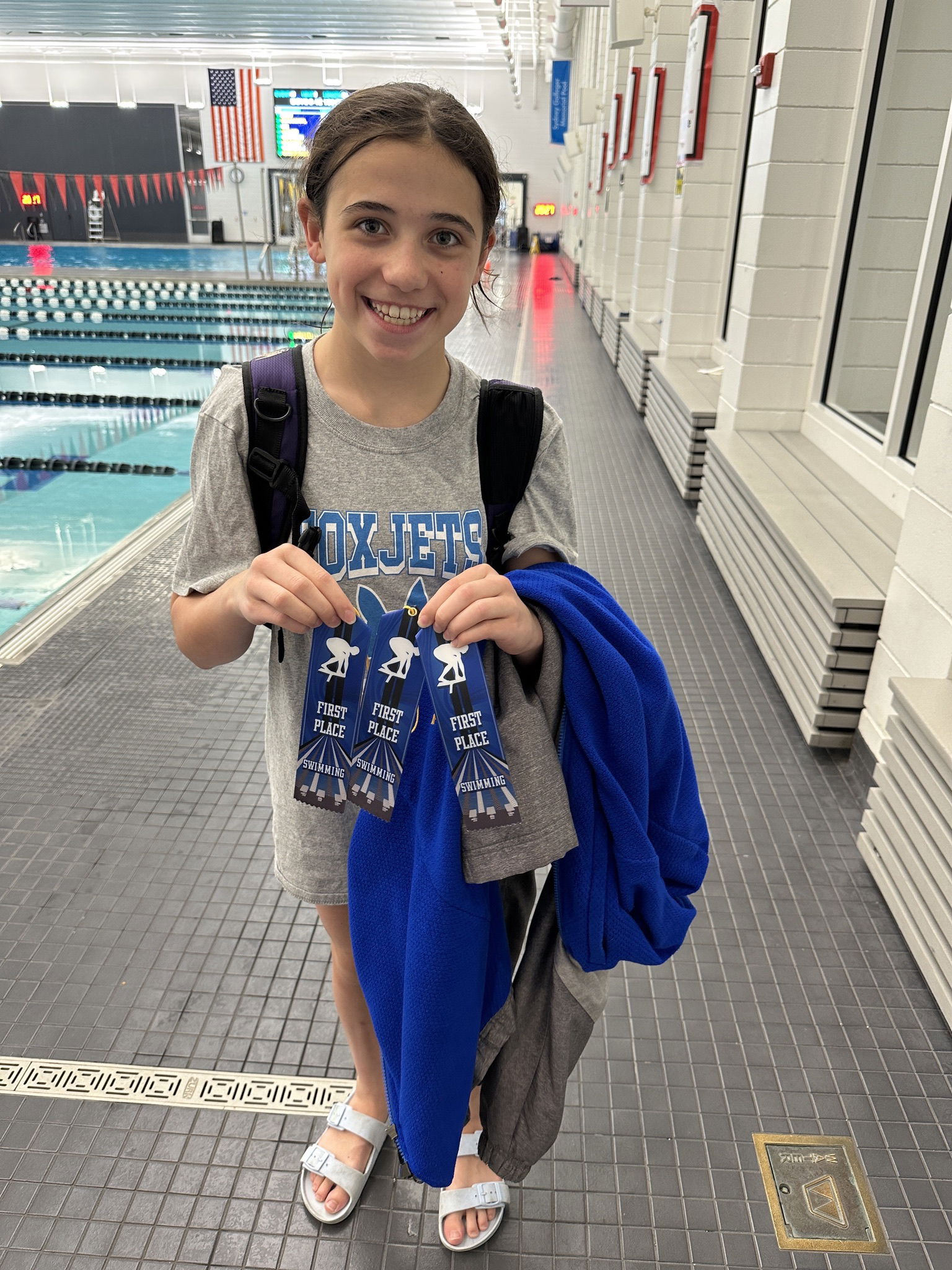 A smiling young girl stands poolside, holding two first place ribbons in swimming. She wears a gray t-shirt, has a backpack slung over one shoulder, and holds a blue towel. In the background, the swimming pool is visible, along with