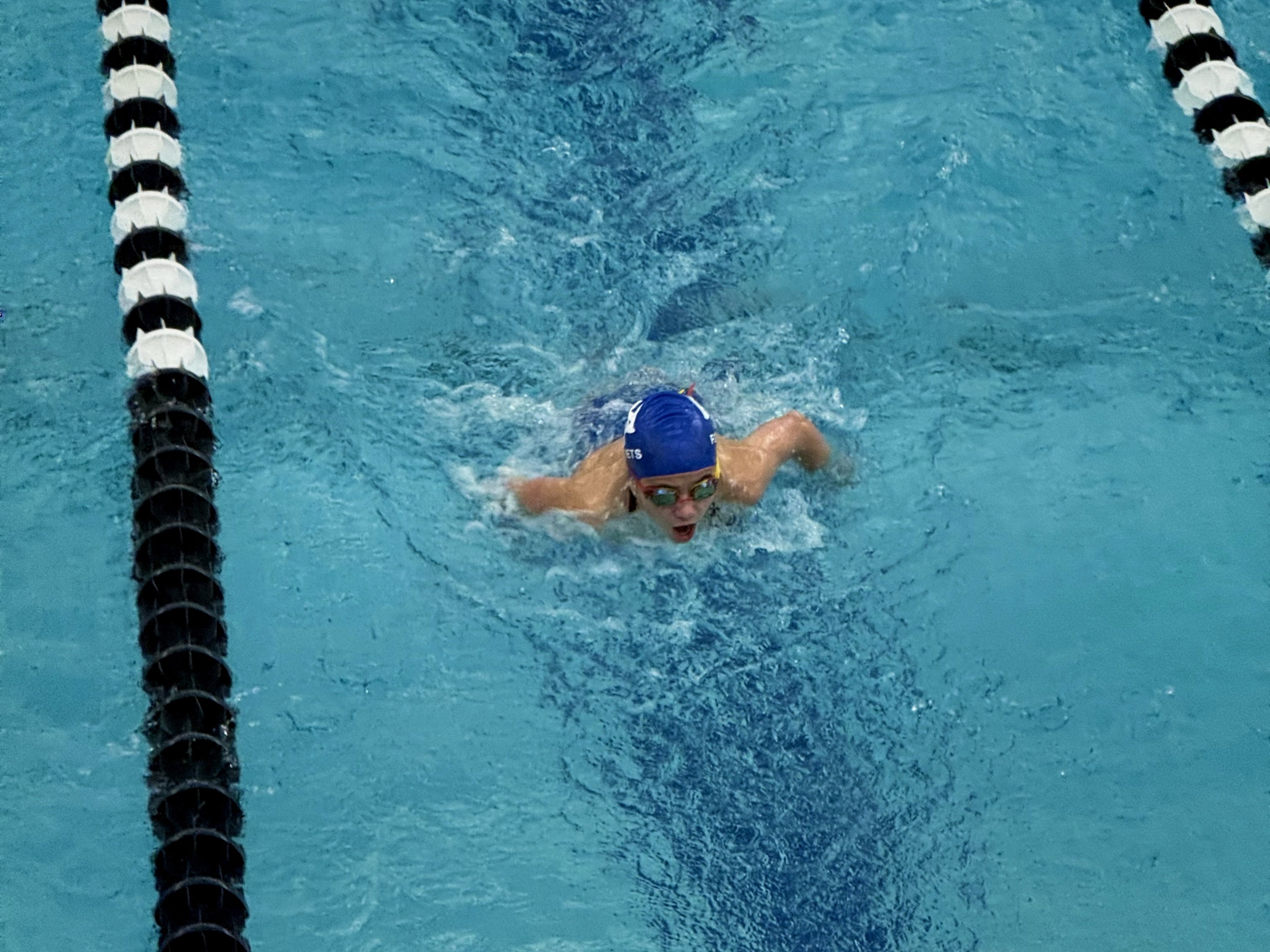 A swimmer in a blue cap and goggles performs a butterfly stroke in a pool, with black and white lane dividers visible alongside.