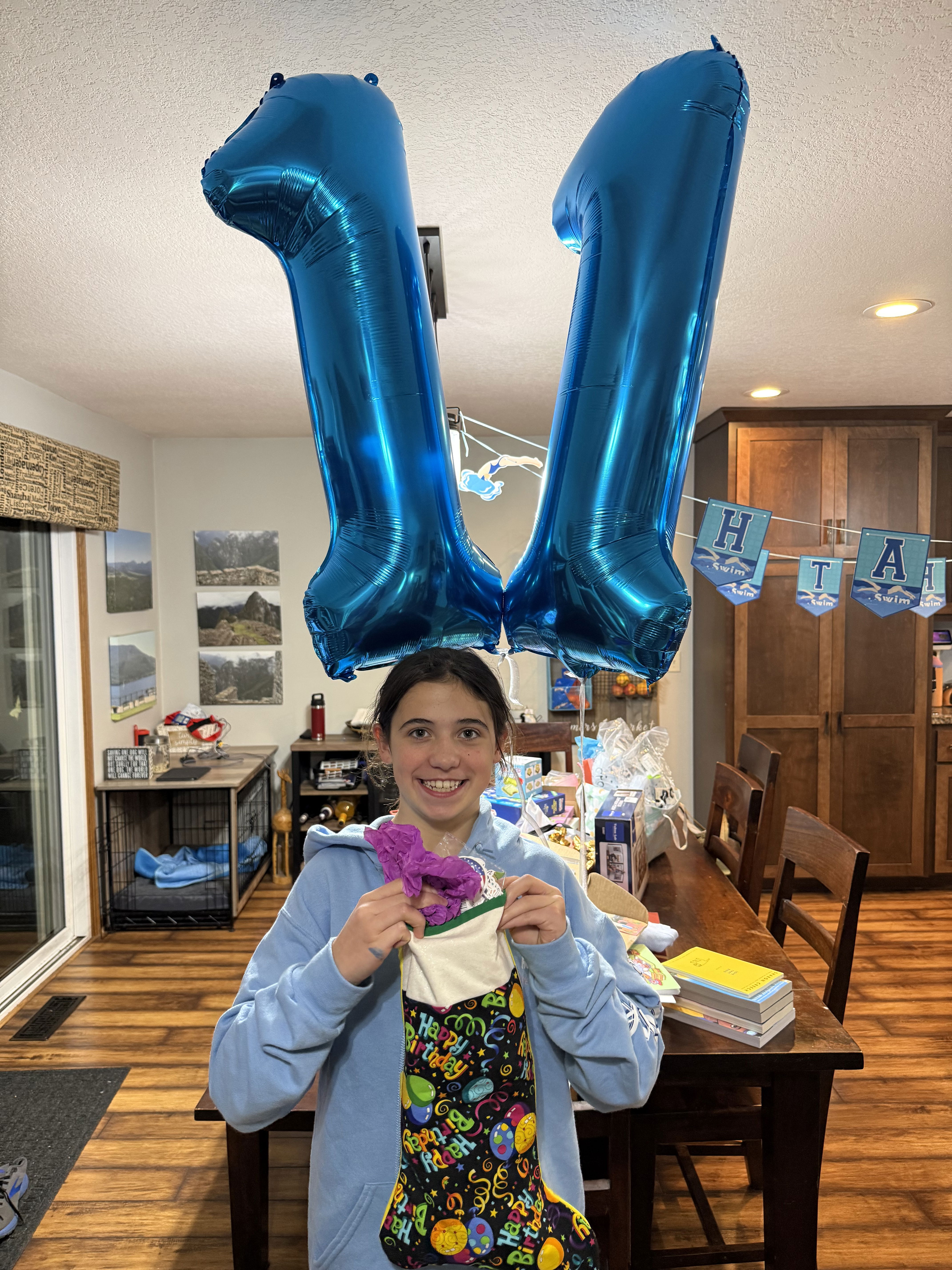 A young girl smiles while holding a colorful birthday stocking and a purple gift inside it. Above her, two large blue balloons shaped like the number 11 float. The setting is a festive room decorated for a birthday celebration.