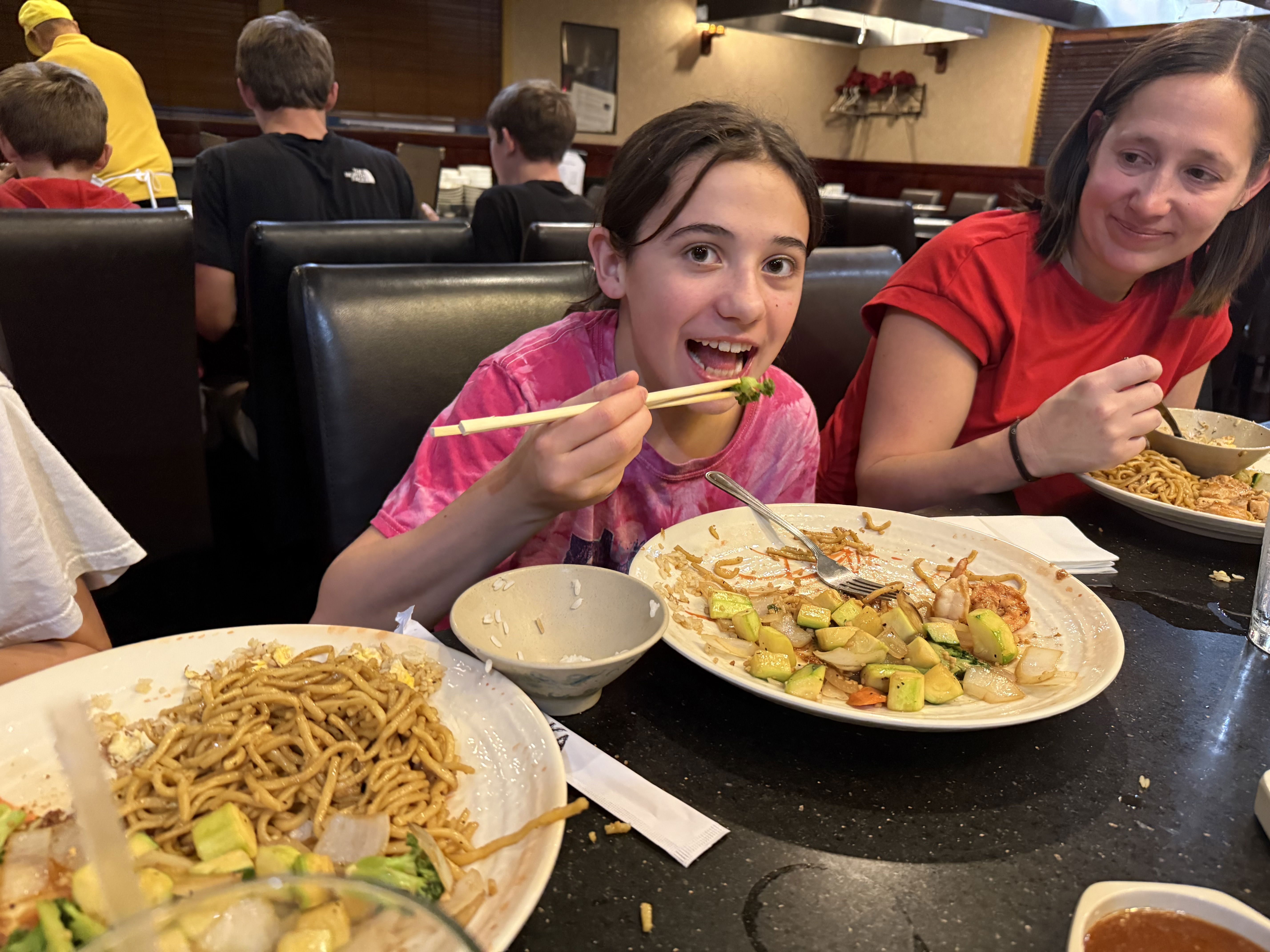 A young girl in a pink tie-dye shirt is smiling and holding chopsticks while eating at a restaurant. She has a plate of stir-fried vegetables and noodles in front of her. A woman in a red shirt sits next to her, also