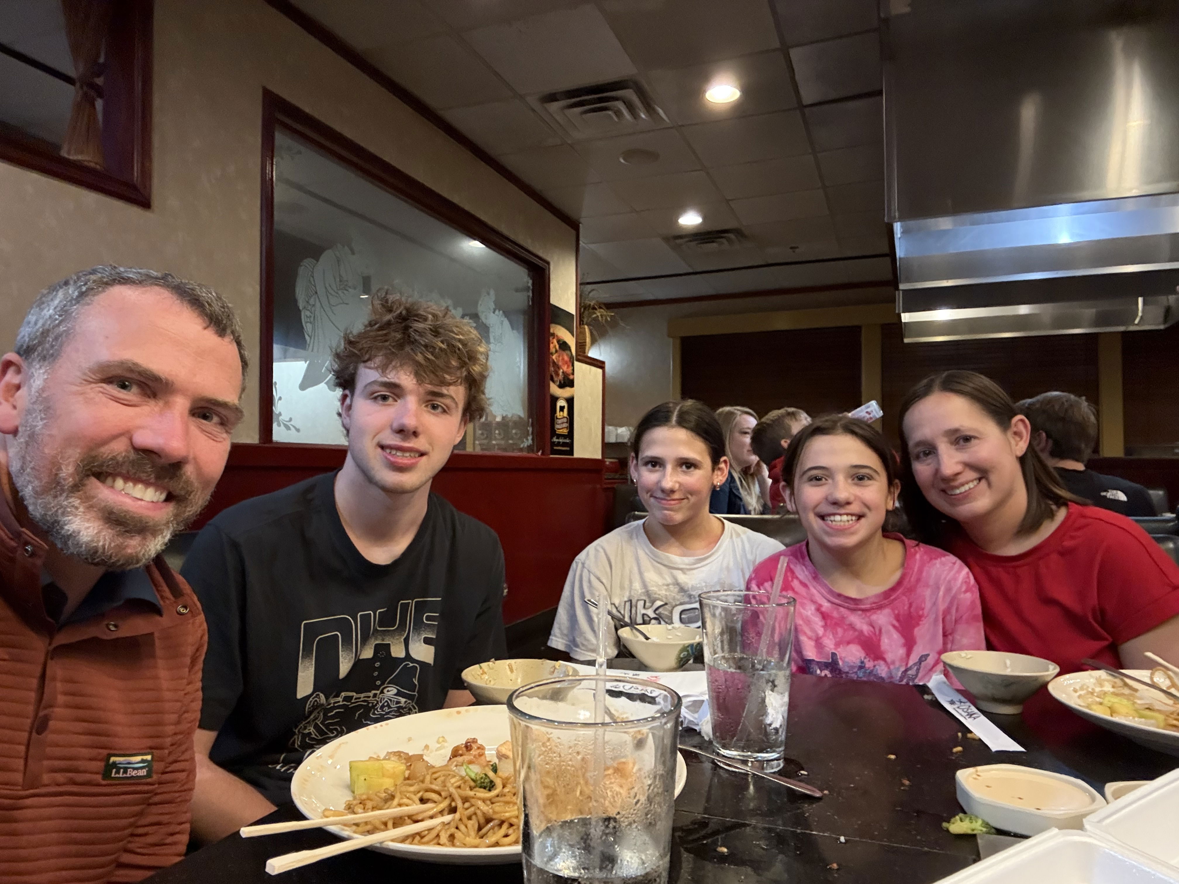A group of five people sits at a restaurant table, smiling for the camera. Plates with food are in front of them, and glasses of water are also on the table. The setting appears casual and lively, with other diners visible in the background.