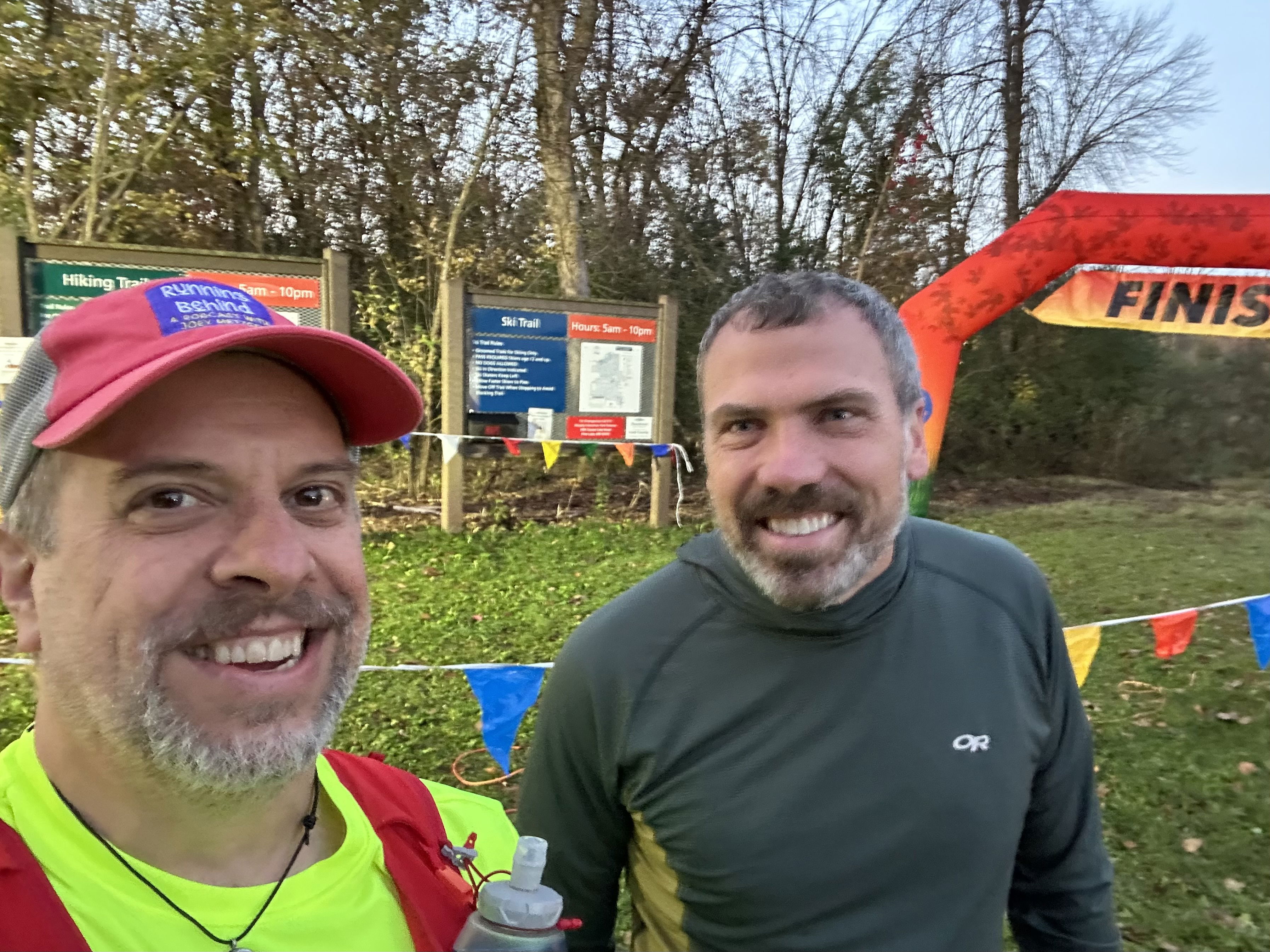 Two smiling men pose together at a race finish line, with a banner reading “FINISH” in the background. They are surrounded by trail signs and colorful flags. One man wears a bright yellow shirt and a red cap, while the other is in
