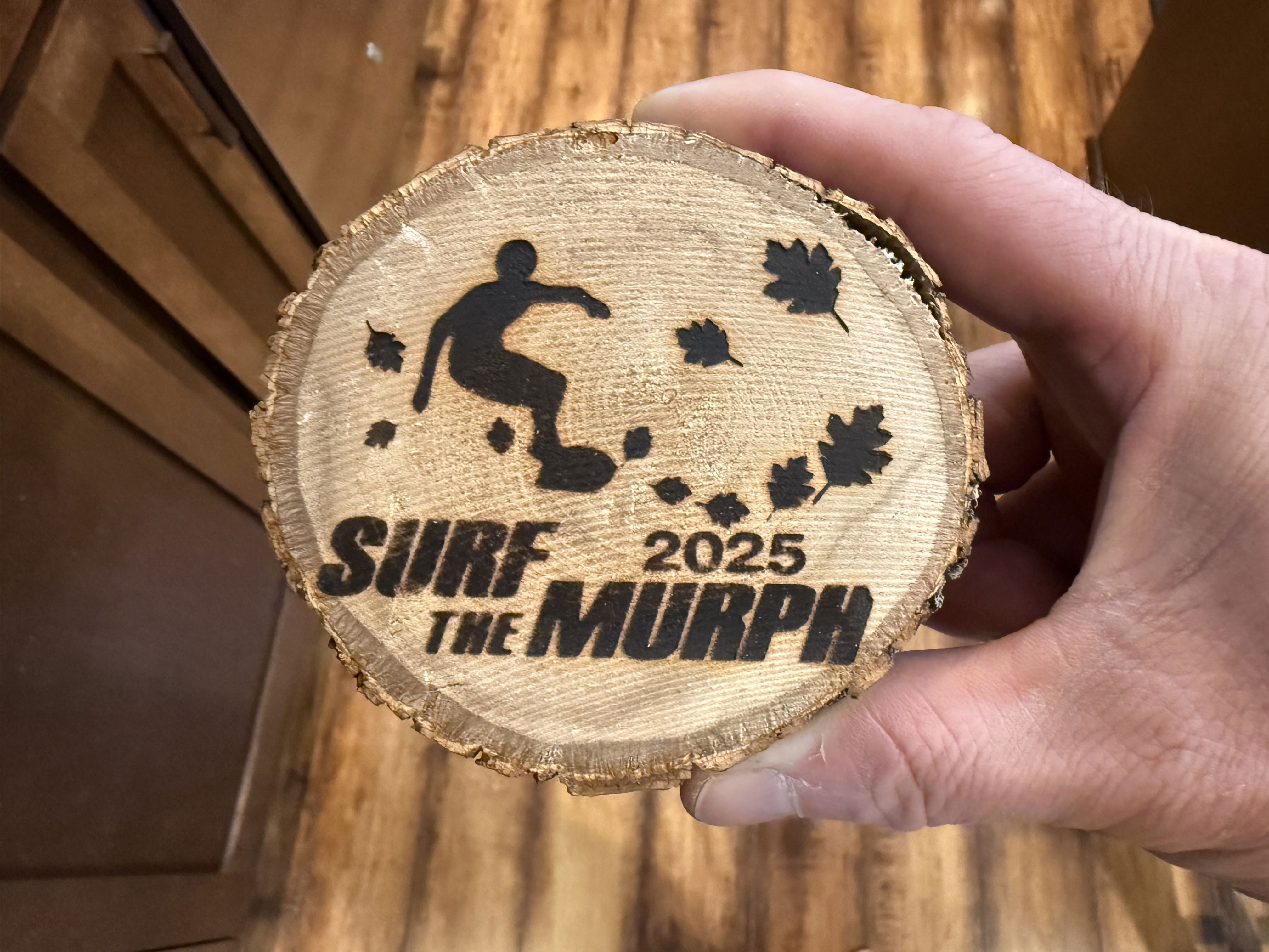 A hand holds a round wooden slice with a black silhouette of a person surfing, surrounded by leaves and the text “SURF THE MURPH 2025.” The background features wooden surfaces.