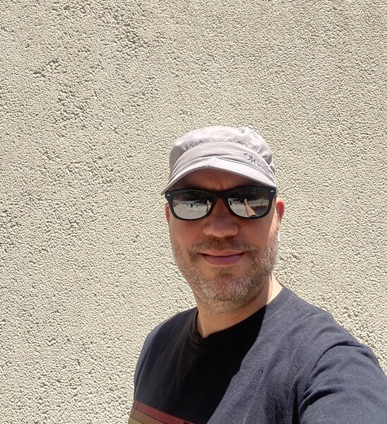 Man in grey baseball hat and black wayfarer sunglasses with a cream colored stucco background 