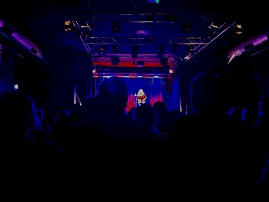 A dark hall in an event center with people sitting on chairs in front of a stage on which a woman sits on a bar stool at a small table to give a reading.