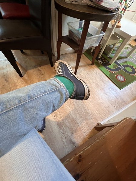 Wazifubo: der Fotograf sitzt auf einer Holzbank und hat ein Bein über das andere geschlagen. Er trägt eine helle Jeans, grüne Socken und blaue Turnschuhe. Der Boden ist helles Laminat, und im Hintergrund ist eine Ecke mit Kinderspielzeug. Dort auf dem Boden liegt ein Verkehrspiel-Teppich.