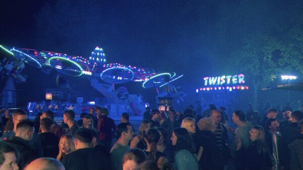 Eine nächtliche Volksfestszene: vor einem beleuchteten Karussell mit Namen 'Twister' steht eine Menschenmenge. Die Menschen unterhalten sich miteinander. Das Bild ist mit einem Blau verstärkenden Filter überlegt.