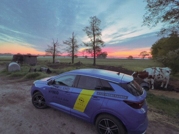 Ein blauer SUV von Opel steht am frühen Morgen am Rande eines Bauernhofes. Auf dem Auto steht Werbung für ein Autohaus.
Der Bauernhof ist nicht zu sehen, aber ein Schuppen, landwirtschaftliche Geräte und eine Kuh.
Am Horizont ist die Morgenröte.