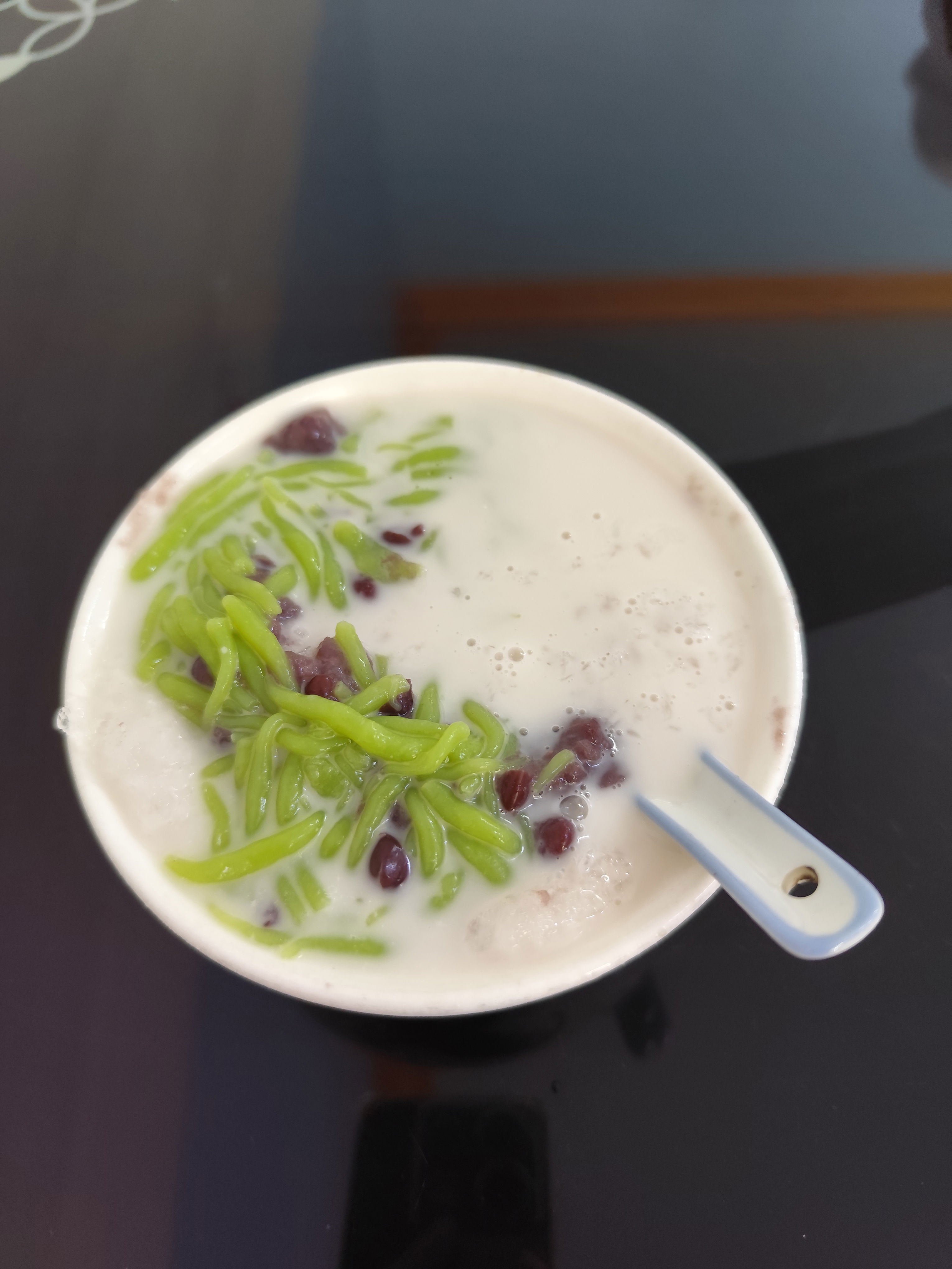 Photo of cendol, an iced sweet dessert that contains pandan-flavoured green rice flour jelly, coconut milk, and palm sugar syrup, in a white bowl with a Chinese spoon.
