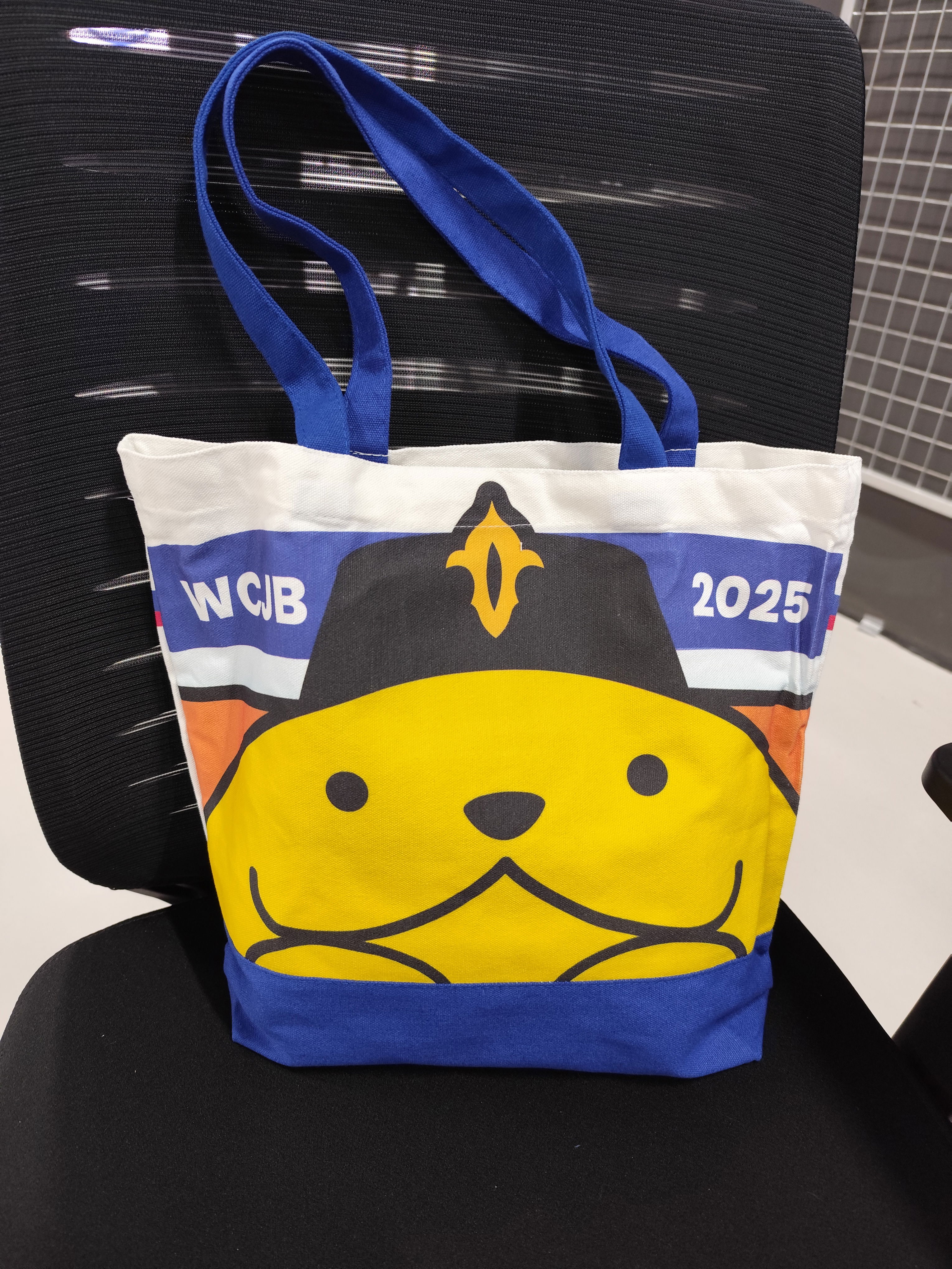 Photo of a tote bag of WCJB 2025, which stands for WordCamp Johor Bahru 2025, with blue handles and the head and hands of the Johor version of Wapuu, the WordPress mascot.