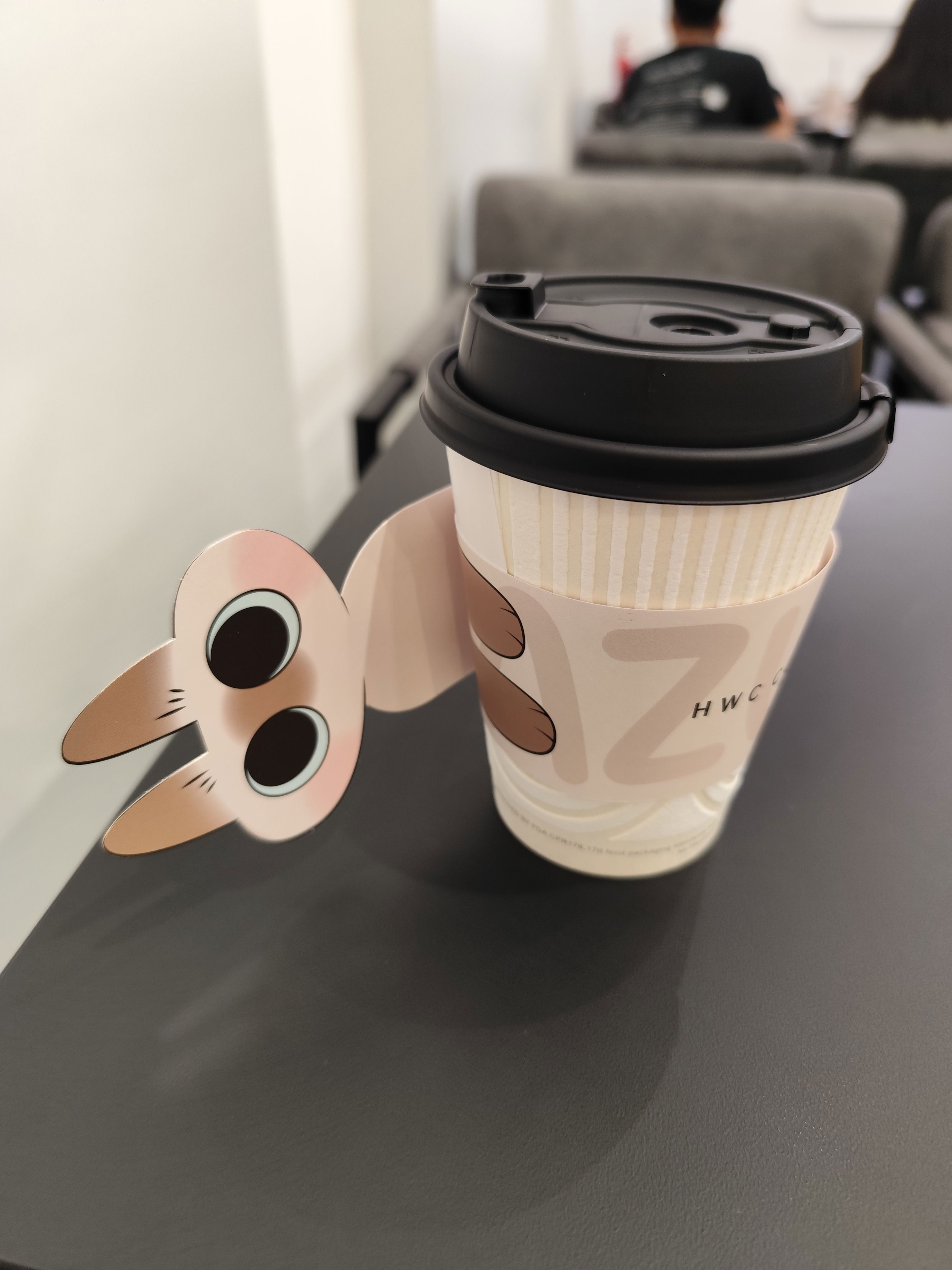Coffee cup of HWC Coffee, with the cup sleeve designed with a cream-coloured cat mascot, appearing as if the cat is wrapping around the cup.