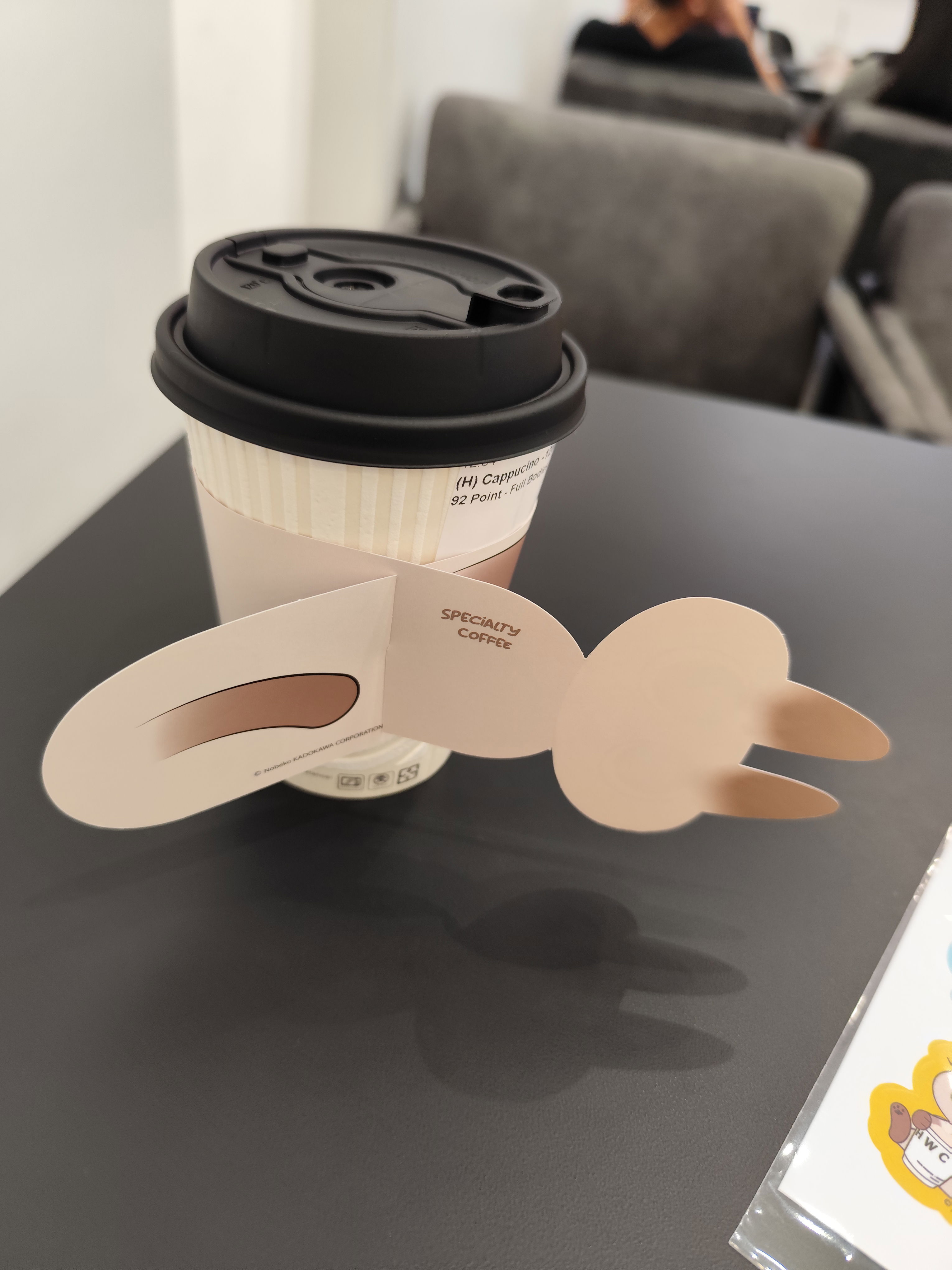Coffee cup of HWC Coffee, with the cup sleeve designed with a cream-coloured cat mascot, appearing as if the cat is wrapping around the cup, while the camera shows the cat mascot's back and tail.