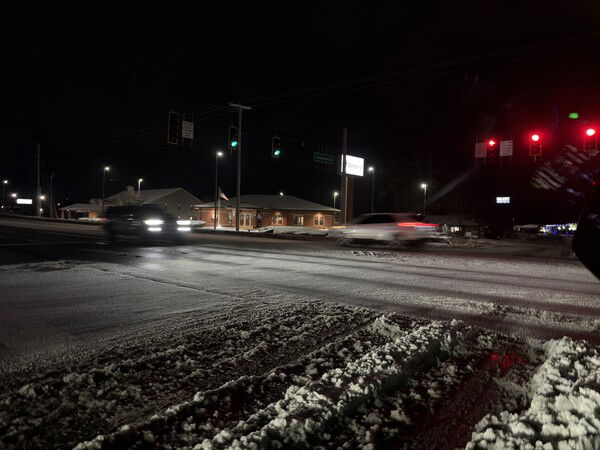 Snowy intersection at night with moving cars, red and green traffic lights, and a building with an American flag under a dark sky.