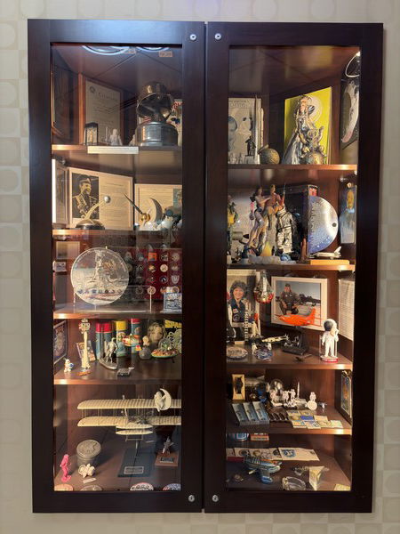Glass cabinet displays aviation memorabilia, including model planes, astronaut figures, certificates, and vintage gadgets, organized on multiple shelves with varied lighting.
