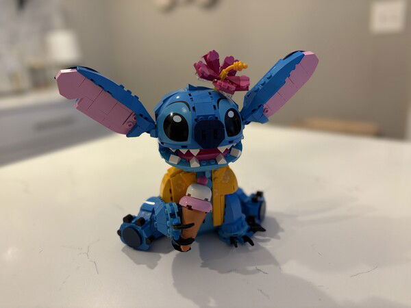  Complete.

Blue LEGO Stitch with large ears, pink inner ears, and a flower on its head, holding a pink ice cream cone, sitting on a white surface.
