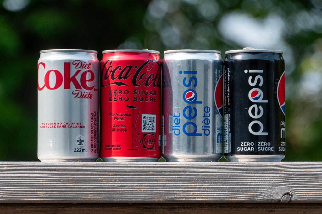 Four 222ml cans of soda on a wood deck rail with blurred foliage in the background. Left to right is a red text on silver Diet Coke, black text on red Coca-Cola Zero Sugar, blue text on silver Diet Pepsi and a white text on black Pepsi Zero Sugar. 
