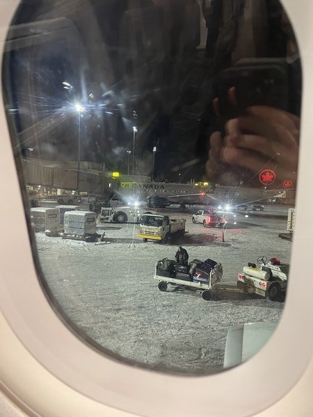 A view out the window of an airplane with a snowy apron and baggage cart in the foreground.