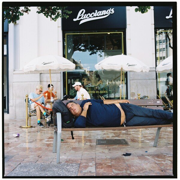 Street photo of a male tourist having a sleep on a bench in front of a fancy looking ice cream shop.