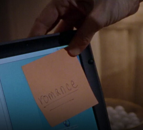The word 'romance' is written on a Post-It note. It's underlined twice. The post-it note is being stuck onto an open laptop's screen in the top right corner.