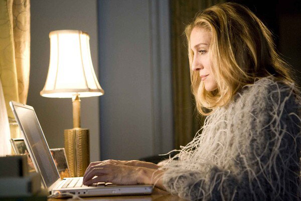 Actress Sarah Jessica Parker, playing the character Carrie Bradshaw from the show 'Sex and the City', types  on a laptop. She is wearing a shaggy fringe jacket, and there's a small lamp on the table illuminating the frame.