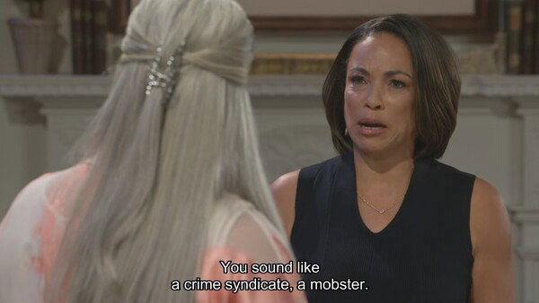 A woman with short dark hair wearing a sleeveless black top looks upset as she speaks to another woman with long gray hair seen from behind. The subtitle at the bottom reads, 'You sound like a crime syndicate, a mobster.'