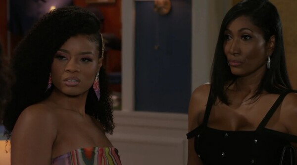 Two women stand indoors looking intently in the same direction. The woman on the left has voluminous curly hair styled to one side, wearing a colorful strapless top and large glittery earrings. The woman on the right has straight shoulder-length hair, wearing a black off-the-shoulder top with thin straps and dangling earrings. Both have serious, slightly skeptical expressions.