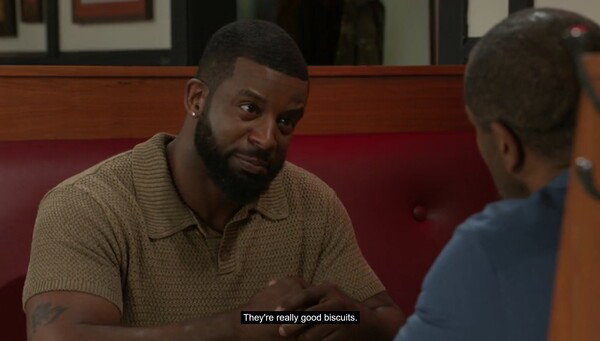 A man with a trimmed beard and short hair, wearing a light brown knit polo shirt, sits in a red booth at a restaurant, clasping his hands on the table and looking intently at another man whose back is to the camera. Subtitles at the bottom read, 'They're really good biscuits.'