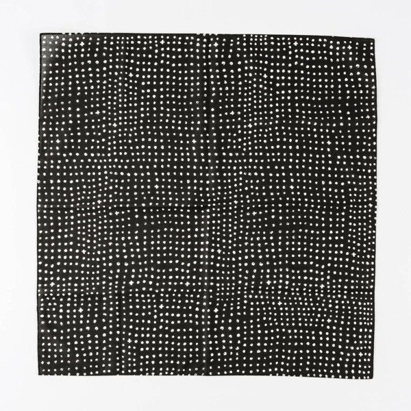 A flat lay photograph of a black square cloth with a white polka-dot pattern. The white dots are irregular in size and spacing, giving the pattern a hand-printed or textured feel. The cloth is folded into a square and placed against a clean, white background, creating a high-contrast, minimalist aesthetic.