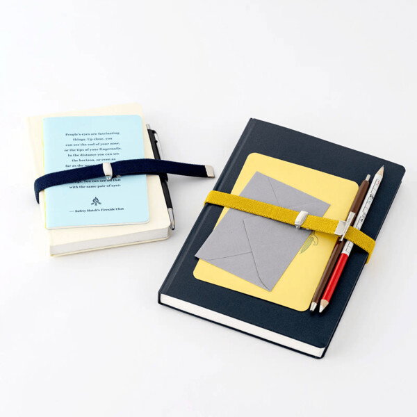 A flat lay photograph showcasing two notebooks secured with adjustable elastic bands. On the left, a cream-colored notebook is bound with a dark blue band, holding a pen. On top of it is a light blue pamphlet with printed text. On the right, a large dark blue notebook is secured with a mustard yellow band, holding two pencils and a gray envelope. The bands have a silver metal clip for adjustment. The image is a clean, bright product shot on a white background, with a focus on organization and accessories.
