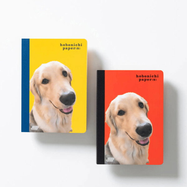A bright product photograph featuring two notebooks with a Golden Retriever puppy's face on the cover. The notebook on the left has a vibrant yellow cover with a blue spine, while the one on the right has a red cover with a black spine. The dog is looking directly at the camera with a happy, smiling expression. The text on both notebooks reads, 'hobonichi paper(S)' with a smaller 'tools & toys' logo on the bottom left. The image has a clean, minimalist style with a solid white background, highlighting the playful and colorful design of the notebooks.