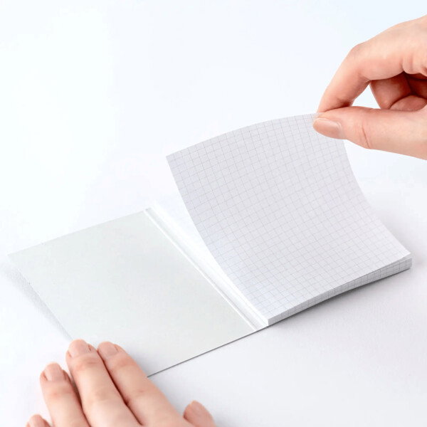 A flat lay photograph showing two hands interacting with a small sticky note pad. The left hand holds open the front cover, while the right hand gently peels back a single sheet of paper from the top of the pad. The paper has a subtle grid pattern on it. The pad and its cover are a clean, crisp white, and the image is shot against a matching white background, creating a bright and clean product-focused aesthetic.