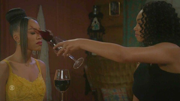 A dramatic screenshot from a television show, capturing a moment of intense confrontation. A Black woman on the right, wearing a black top with a sleeveless cut, is captured mid-motion as she pours a glass of red wine onto the face of another Black woman on the left. The woman being poured on has her eyes closed and is wearing a bright yellow, lace-trimmed top with a thin gold necklace. Her hair is styled in a bun with two braids framing her face. A full glass of wine sits untouched on the table below. The image has a high-tension, shocking, and emotionally charged mood.
