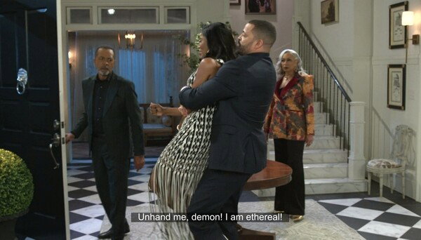 A tense and dramatic scene from a television show, set in a grand, black-and-white checkered foyer. A bald white man in a dark suit is holding a Black woman in a black and white patterned dress with a fringed skirt, lifting her off the ground as she struggles. Another Black man in a dark suit stands to the left, holding a door open with a surprised expression. A white-haired woman in a colorful, patterned jacket stands to the right by a staircase, looking on with concern. Subtitles at the bottom read, 'Unhand me, demon! I am ethereal!' The image captures a highly theatrical and chaotic moment.