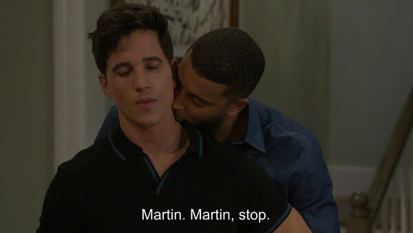 A television screenshot from a dramatic scene showing a white man in a black polo shirt with his eyes closed and a neutral expression, as a Black man embraces him from behind and kisses his cheek. The Black man is wearing a blue button-up shirt. The subtitle at the bottom reads, 'Martin. Martin, stop.' The scene has a tense and emotional mood.