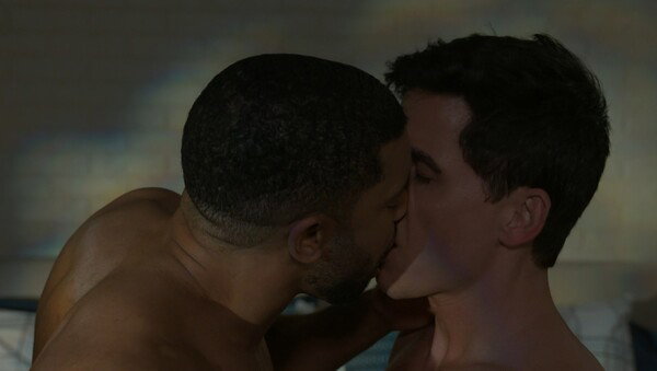 A close-up, dimly lit screenshot showing two men in profile during a romantic moment. A Black man, whose face is partially obscured and is bare-chested, is passionately kissing a white man with dark hair, who is also partially bare-chested. The mood is intimate and romantic.