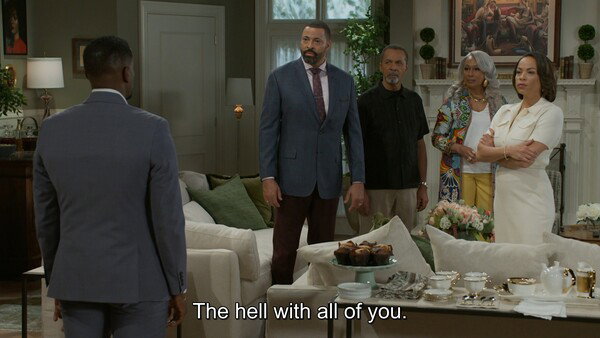 A wide shot from a television show set in a lavishly decorated living room with an off-white sofa and coffee table. A man in a suit stands with his back to the camera, facing a group of four people. The group consists of a bearded Black man in a blue suit, an older man in a black shirt, a woman with gray hair in a colorful jacket, and a woman in a white dress with her arms crossed. The subtitle at the bottom of the screen reads, 'The hell with all of you.' The mood is tense and dramatic.