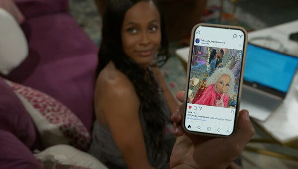 A candid shot shows a person holding a smartphone, with a social media post clearly visible on the screen. The post features a photo of an older woman with vibrant white hair, wearing a bright pink suit and making a humorous face, possibly from a wedding or party. The person holding the phone is slightly out of focus in the background, smiling faintly. This image captures a moment of sharing content with someone nearby.