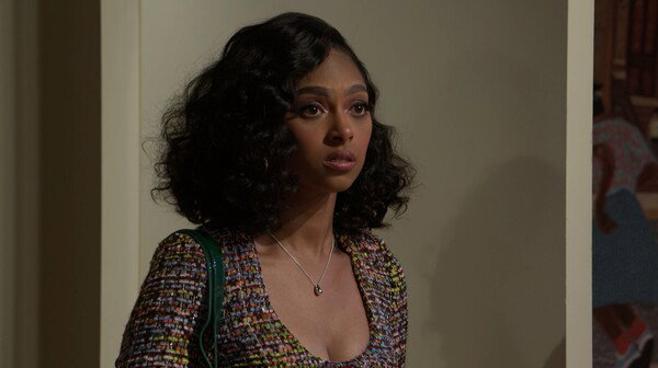 A medium-close shot of a Black woman with voluminous, dark, curly hair looking off-camera with a surprised or concerned expression. She wears a multi-colored tweed-style blazer or jacket with a low-cut neckline and a small, silver pendant necklace. The background is an interior setting with neutral-toned walls, and a hint of a patterned object (possibly a quilt or tapestry) is visible on the far right. The image has the bright, well-lit, and polished aesthetic of a television drama or sitcom.