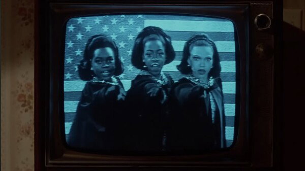 A low-resolution image of a vintage television screen displays a scene in monochrome with a strong blue tint. Three young Black women with elaborate hairstyles are standing side-by-side, wearing dark capes or robes and extending their right arms with their fingers pointing forward. They are positioned in front of a large American flag backdrop. The image is captured within the wooden casing of an old television set, emphasizing a retro or archival context. The overall mood is intense and dramatic, conveying a performance with a powerful, stylized presentation.