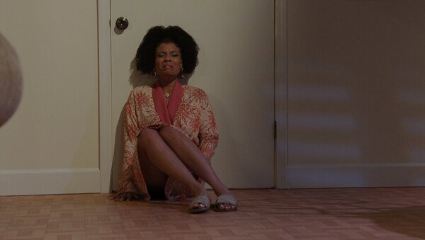 A woman with a large afro hairstyle and distressed facial expression is sitting on a parquet floor, leaning against a white closed door. She is wearing an open, patterned robe in shades of coral and cream, with a glimpse of a pink lining visible, and simple slide sandals. Her legs are crossed and exposed. The lighting is dim and warm, creating a dramatic mood that suggests a moment of intense emotion or despair in a film or theatrical setting.