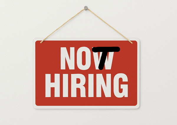 A high-contrast image featuring a red rectangular sign hanging on a plain white wall by a thin piece of twine. The sign has the large white text 'NOW HIRING'. The word 'NOW' has a thick, black horizontal line crudely drawn over the letter 'N' and part of the 'O', changing the meaning to imply 'NOT HIRING'. The style is simple and graphic, with bright colors and a clear, ironic context about a change in employment status.