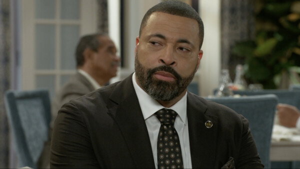 A close-up of a middle-aged Black man with a well-groomed salt-and-pepper beard, wearing a dark brown or black suit, a white dress shirt, and a dark tie with a subtle pattern. He is seated at a table in what appears to be a formal restaurant or dining room, looking off to the side with a serious, slightly intense or contemplative expression. In the blurred background, another man in a suit is visible, suggesting a business or formal meeting context. The image has a warm, indoor lighting style, typical of a dramatic scene from a television show or movie.