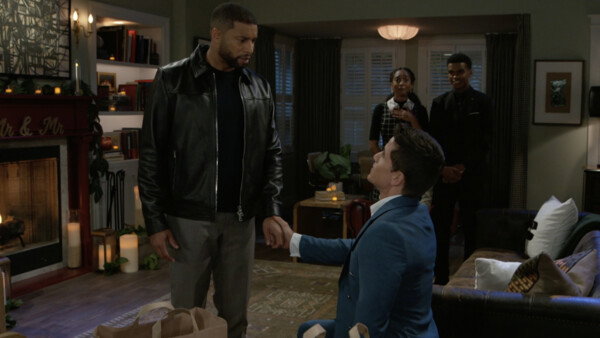 A scene depicting a marriage proposal indoors. A light-skinned man wearing a teal blue suit is down on one knee, looking up at and holding the hand of a dark-skinned man in a black leather jacket and gray pants. The man in the jacket looks down with a slightly shocked or emotional expression. Two young Black people—a woman in a plaid dress and a man in a black suit—stand behind them in the mid-ground, observing the moment. The room is decorated with a warm, romantic atmosphere, featuring numerous lit candles, a fireplace, and a sign that reads 'Mr. & Mr.' over the mantel. The overall style is that of a contemporary television comedy-drama.
