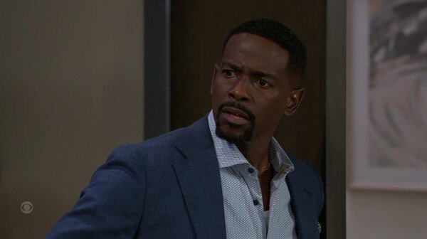 A medium-close-up shot shows a Black man in a navy blue suit jacket and an unbuttoned, patterned light blue collared shirt. He has a neatly trimmed beard and mustache. He is looking sharply to his left with a concerned, surprised, or intense expression, his eyebrows slightly furrowed and his mouth slightly open. The background is indoors, mostly blurred, showing a dark wall or doorway on the left and a framed picture on the right. The image has the high-definition and dramatic lighting typical of a television drama or soap opera still, suggesting a moment of high tension or revelation.