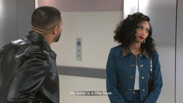 A still from a television show shows a Black man and woman standing in front of two brushed metal elevator doors in a modern interior. The man, on the left, has a close haircut and is wearing a black leather jacket; he is looking towards the woman, but his face is mostly turned away from the camera. The woman, on the right, has voluminous dark, curly hair and is wearing a blue denim jacket with a brown leather collar and gold buttons, along with matching denim bottoms. She is looking slightly off-camera to her right with a serious or mildly annoyed expression and has bright red lipstick. A white subtitle across the bottom of the screen reads: 'My sister is a little freak.'
