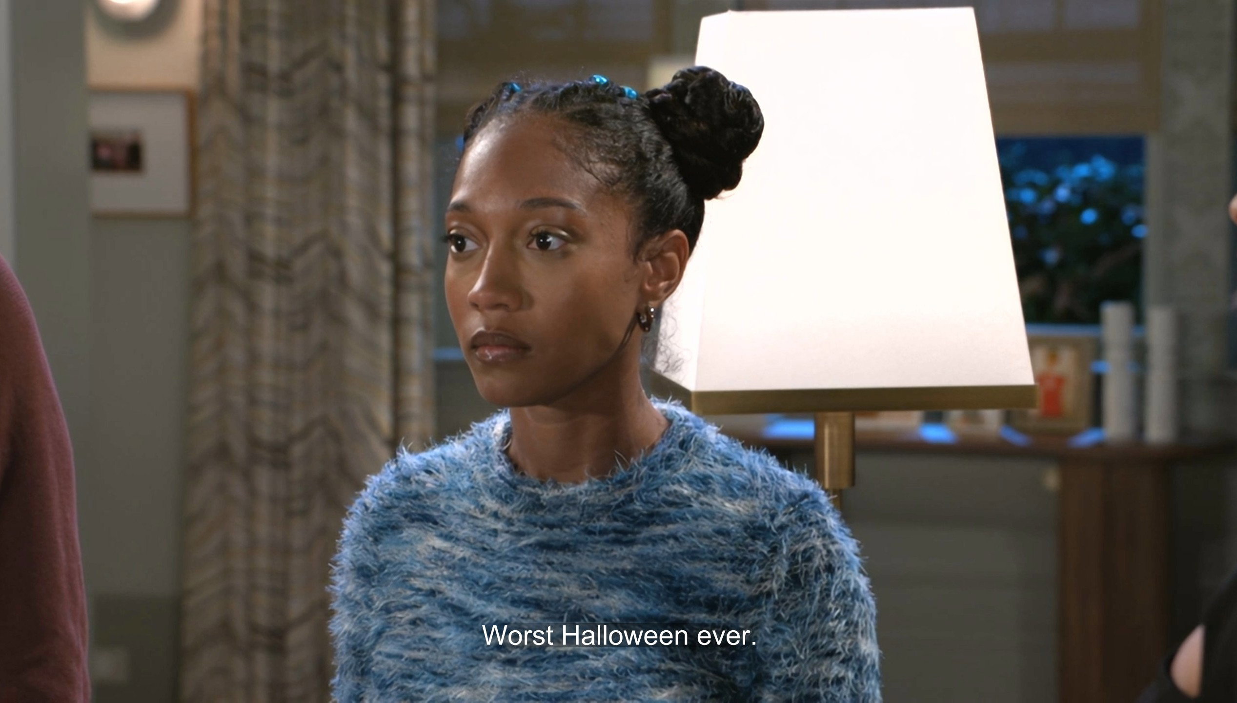 A medium shot of a young Black woman with her hair in a high braided bun and wearing a textured blue and white fuzzy sweater. She looks off-camera with a serious or disappointed expression. Below her, a subtitle reads, 'Worst Halloween ever.' The scene is an interior with soft, professional television lighting, suggesting a dramatic or comedic moment from a TV show or film.