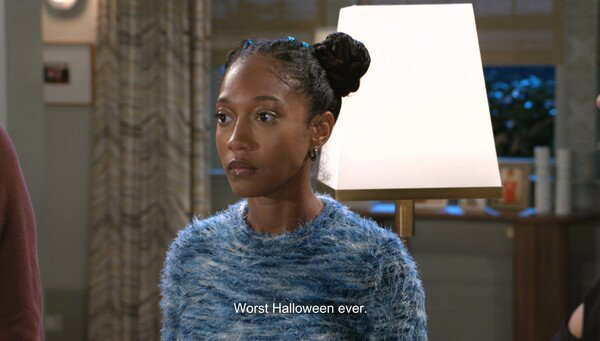A medium shot of a young Black woman with her hair in a high braided bun and wearing a textured blue and white fuzzy sweater. She looks off-camera with a serious or disappointed expression. Below her, a subtitle reads, 'Worst Halloween ever.' The scene is an interior with soft, professional television lighting, suggesting a dramatic or comedic moment from a TV show or film.