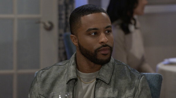 A close-up shot of a young Black man with a close-cut hairstyle and a neatly trimmed beard, looking intently off-camera to the right. He is wearing a silver or light grey distressed leather-look jacket over a light-colored t-shirt and a thin gold chain. The background is softly blurred, showing an indoor setting that appears to be a dining area with a glass-paneled door and another person faintly visible. The overall mood is serious or contemplative, suggesting a scene from a television show or film.